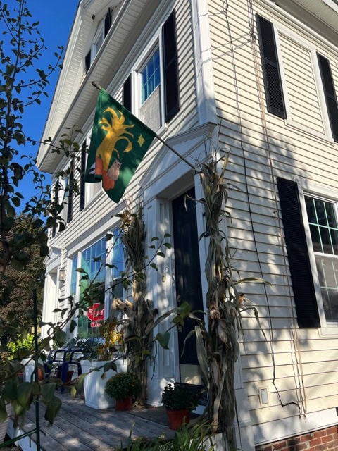 a two story white house with black shutters, a green flag featuring a rooster, cornstalks by the door, and potted plants on a wooden porch.