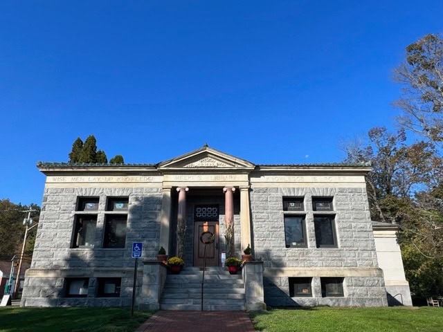 a small historic stone library building with large windows, a central entrance with columns, and a clear blue sky in the background.