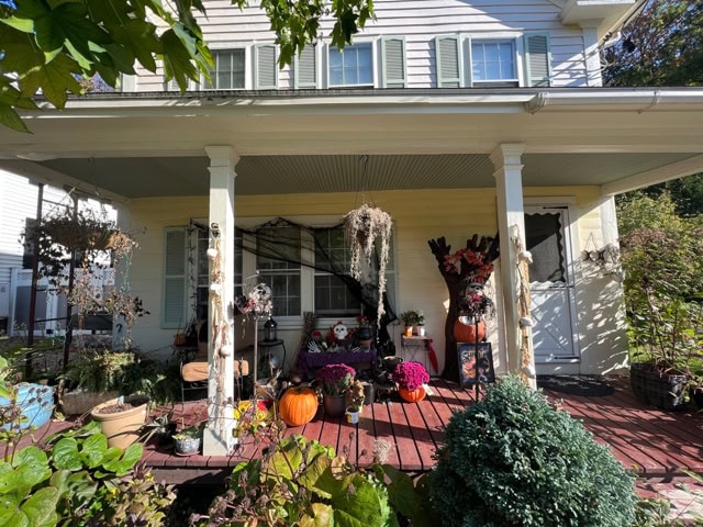 a front porch decorated with pumpkins, mums, scarecrows, and hanging plants, suggesting a fall or halloween theme. the house has light siding and green shutters.