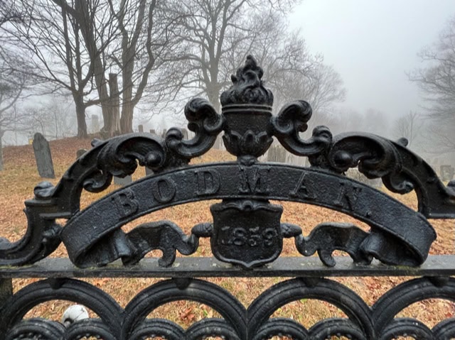 ornate black iron cemetery gate with "bodman" and "1859" inscribed; tombstones and bare trees in a foggy background.