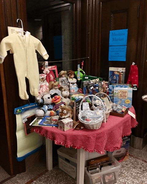 a small indoor market table with toys, board games, dolls, books, and a yellow baby onesie on display, under a sign reading "meekins market.