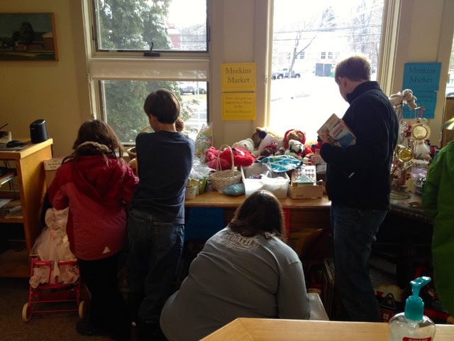 four people look at toys and stuffed animals arranged on a table by a window in a room, with signs reading "meekins market" visible on the wall.