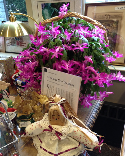 a basket with blooming pink christmas cactus sits on a table with a handmade doll, holiday decorations, and a framed article titled "creating a new tradition.