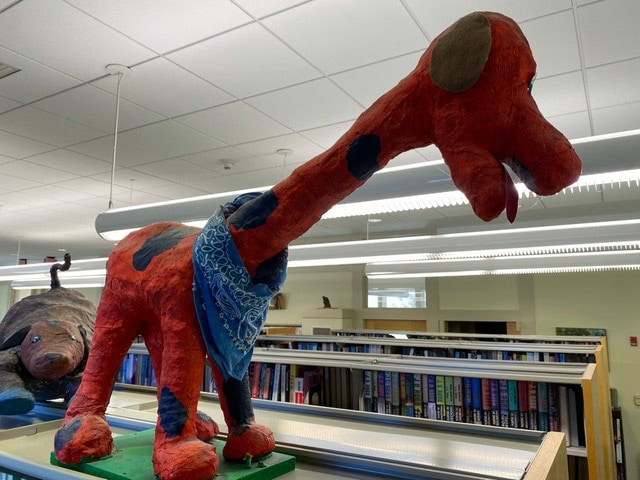 a large red papier mâché dinosaur with blue spots and a blue bandana stands on top of a bookshelf in a library.