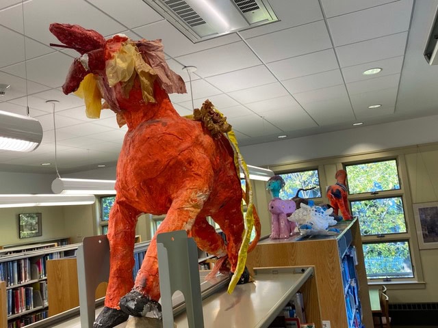 a large red orange papier mâché unicorn with yellow paper mane is displayed on top of bookshelves in a library, with other colorful sculptures visible in the background.