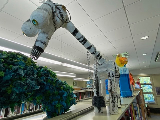 a papier mâché giraffe sculpture with black stripes and a chain around its neck stands on top of bookshelves in a library.