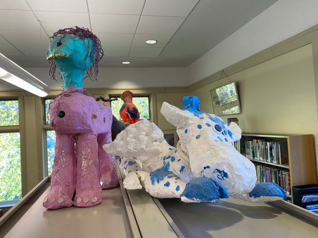 colorful papier mâché animal sculptures, including one purple and one blue and white, are displayed on top of a shelf in a room with windows and bookshelves.