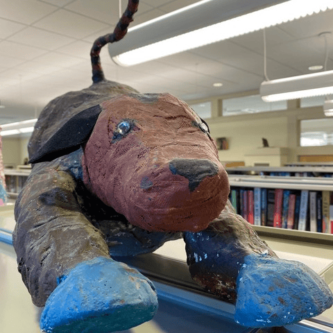 a sculpture of a dog with blue paws and a brown body is displayed on top of a bookshelf in a library, with rows of books visible in the background.
