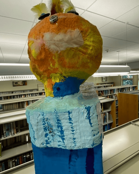 large, colorful papier mâché sculpture with an orange head and blue and white body stands in a library, surrounded by bookshelves and fluorescent lights.
