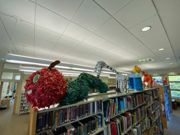 large colorful caterpillar sculptures sit atop bookshelves in a library, with rows of books and windows visible in the background.