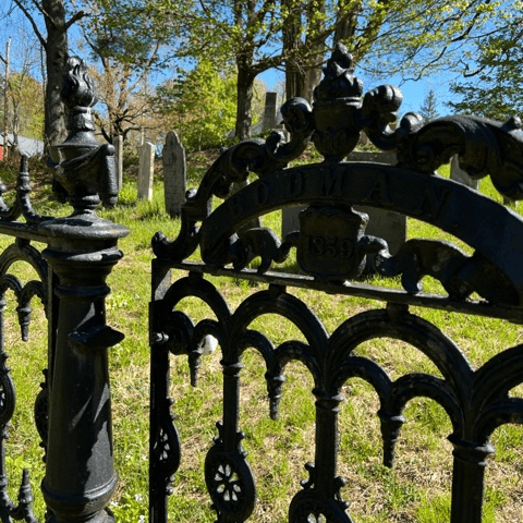 ornate black iron cemetery gate with "goodman 1839" inscribed, set in a grassy graveyard with tombstones and trees in the background under a blue sky.