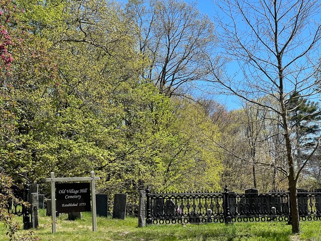 a cemetery with a black iron fence and tombstones is surrounded by trees. a sign reads, “old village hill cemetery established 1771.”.