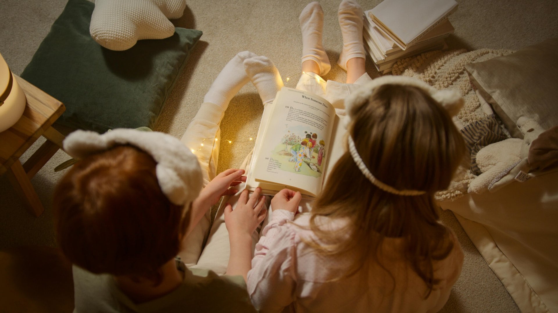 two children wearing fuzzy headbands sit on the floor under a blanket, reading an illustrated book together, lit by string lights.