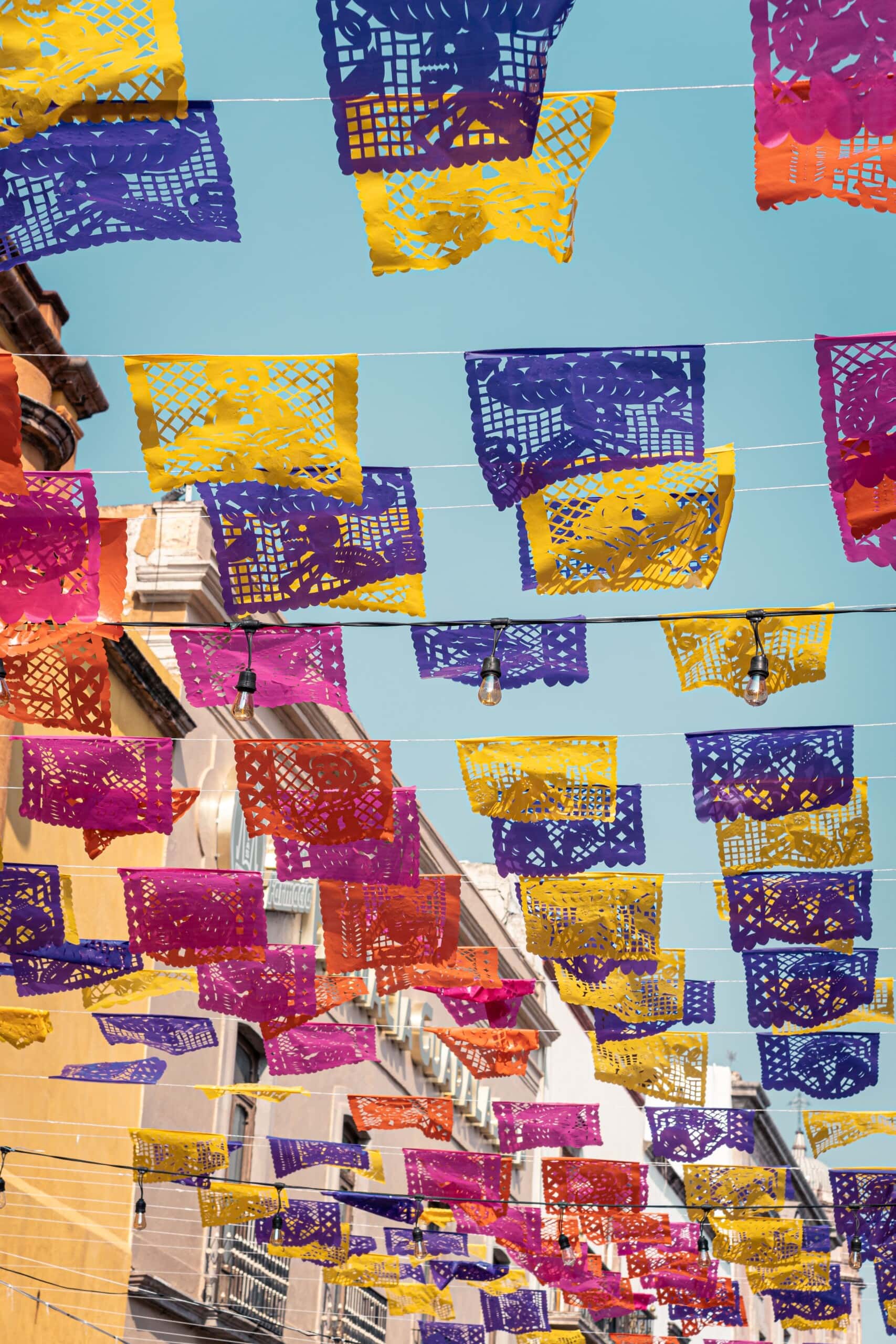 colorful papel picado banners in purple, yellow, and red hang across a street with buildings and balconies visible under a clear blue sky.