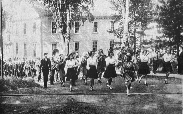 a black and white photo of a marching band with uniformed musicians and baton twirlers walking on a street beside a large building and trees.
