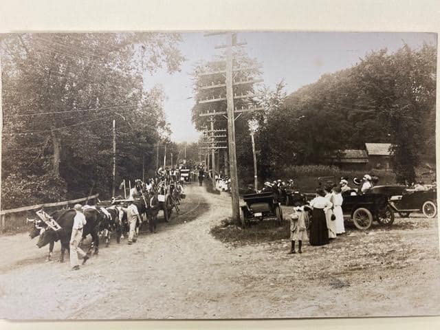 old photograph showing a dirt road with horse drawn wagons and early automobiles, people standing nearby, and trees in the background.