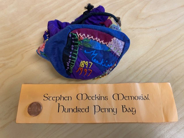 a colorful drawstring bag labeled "stephen meekins memorial hundred penny bag" sits on a table next to a penny and a printed sign.