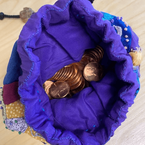 a small fabric drawstring pouch with a purple lining, partially open to reveal several pennies inside.