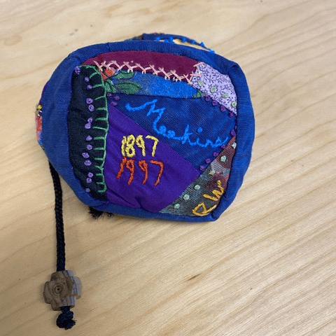 a small, colorful patchwork pouch with embroidery reading "my mending 1897 1997" sits on a wooden surface. a black cord with a bead is attached.