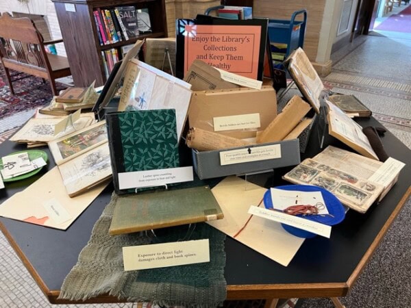 a library display table shows old books, textiles, and signs about preservation, including warnings about light damage and handling best practices.