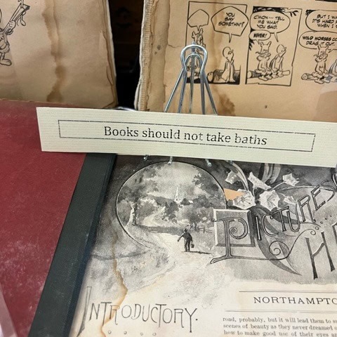 a paper sign reading "books should not take baths" is clipped to a display above old, water damaged books and papers.