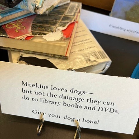a sign reads, "meekins loves dogs—but not the damage they can do to library books and dvds. give your dog a bone!" chewed books and cases are visible behind the sign.