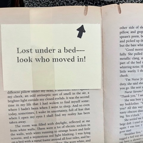 a card reading "lost under a bed look who moved in!" with an arrow is clipped to the top of an open book.