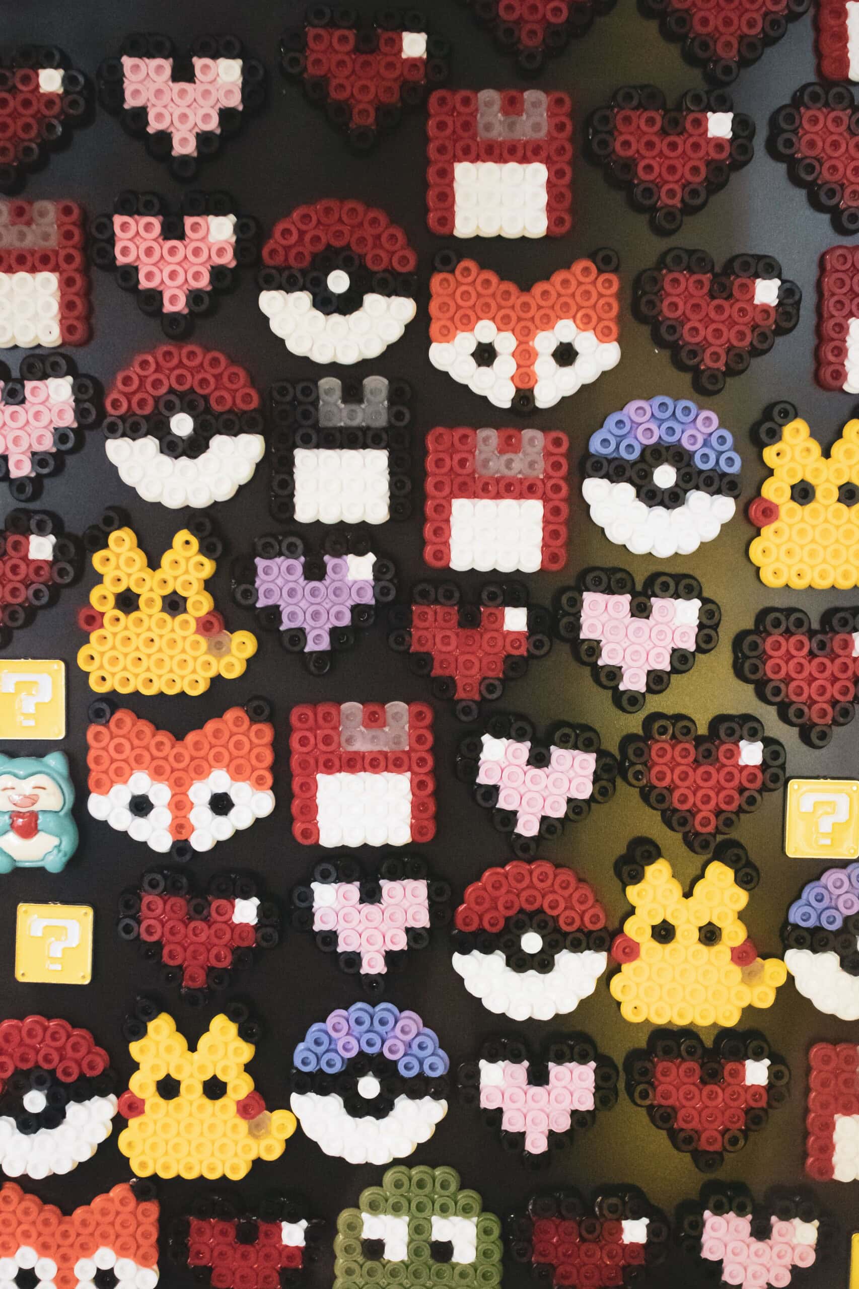 assorted pixel art designs made from fused beads, including poké balls, hearts, fox faces, crowns, and question mark boxes, arranged on a black background.
