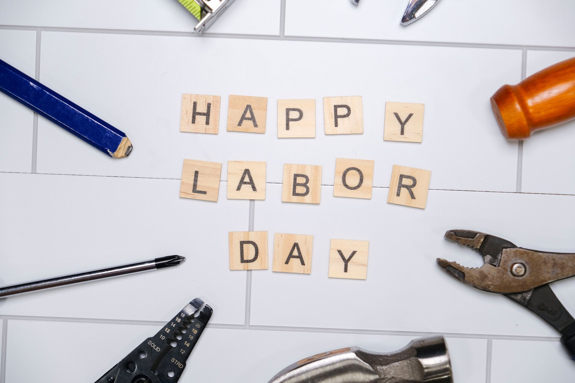 scrabble tiles spelling “happy labor day” are arranged on a white surface, surrounded by various tools including pliers, a screwdriver, and a wrench.