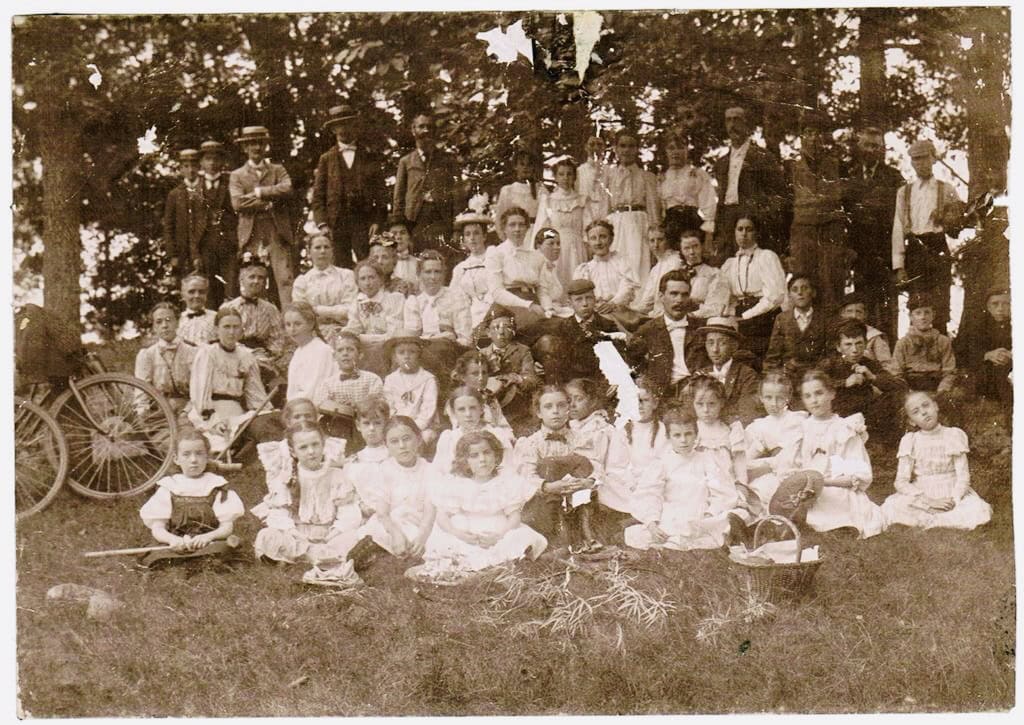 a large group of men, women, and children posed outdoors, sitting and standing in front of trees, with picnic baskets and a bicycle visible.