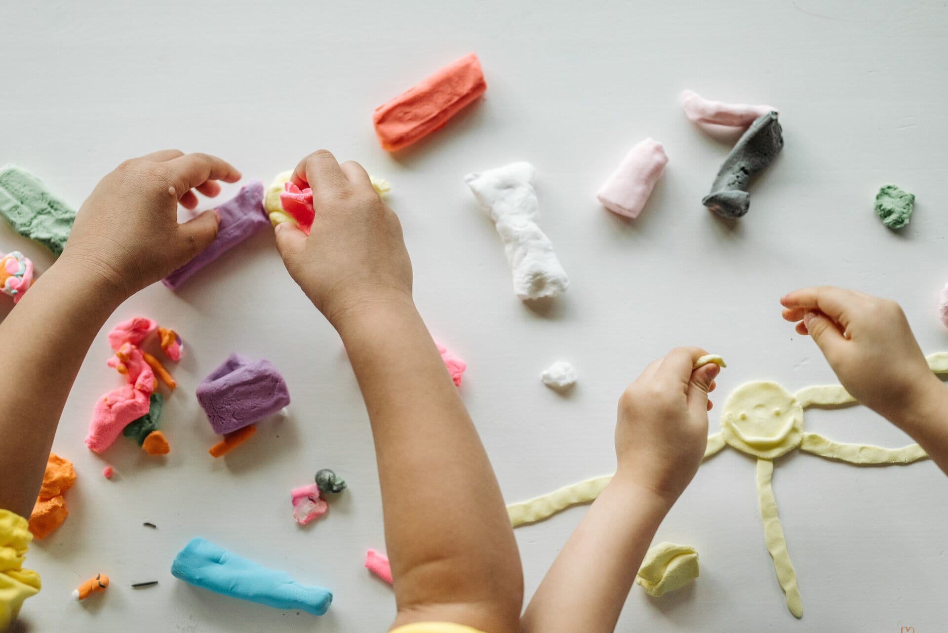 three children’s hands play with colorful modeling clay on a white surface, shaping various pieces and figures.