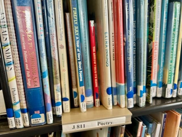 a close up of a library shelf labeled "j 811 poetry," displaying various children's poetry books arranged vertically.