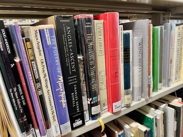 a library bookshelf displays poetry books by various authors, including maya angelou, john ashbery, and grace schulman, arranged upright in a single row.