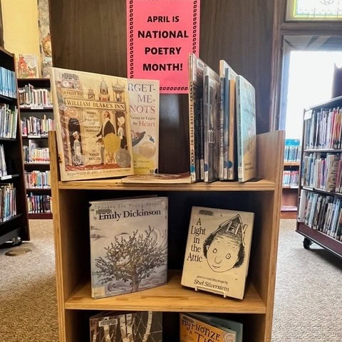 a library book display features poetry books with a pink sign above that reads, "april is national poetry month!" shelves and windows are visible in the background.
