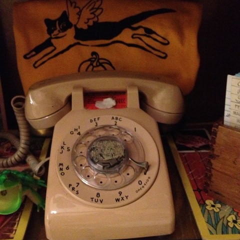 beige rotary dial telephone in front of a yellow cloth with a drawing of a winged cat. various small items are partially visible around the phone.