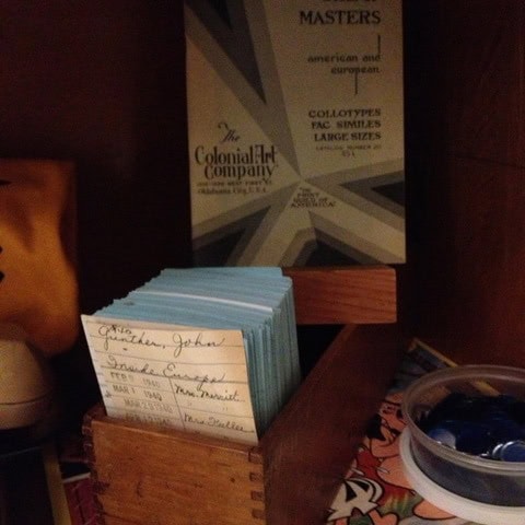 a wooden box holds index cards with handwritten notes. behind it is a colonial art company masters print set. a partial container with blue game pieces is also visible.