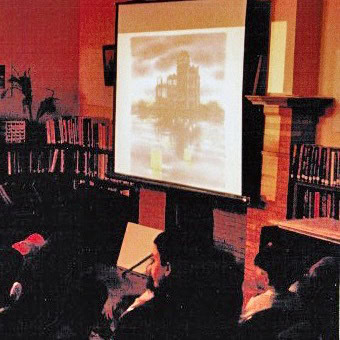 a small group of people sit in a dimly lit room, watching a projection of a building on a screen in front of bookshelves.