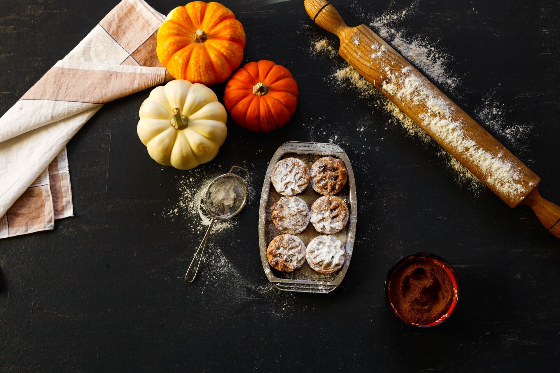 three orange pumpkins, one white pumpkin, a cloth, a rolling pin with flour, a glass dish of six small pastries, a sifter, and a bowl with brown powder on a dark surface.