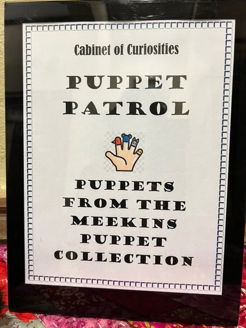 framed sign reads "cabinet of curiosities: puppet patrol—puppets from the meekins puppet collection," with an illustration of a hand wearing finger puppets.