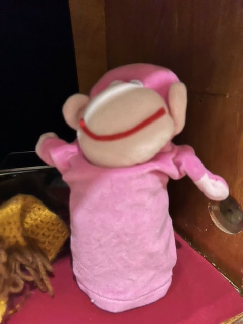 a pink sock monkey puppet with a white face and red mouth is positioned facing away on a shelf next to a yellow crocheted object.