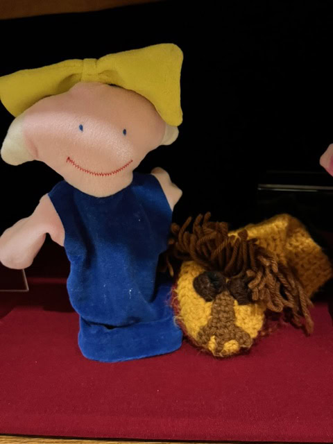 a cloth hand puppet with a yellow bow and blue dress sits next to a knitted lion with a brown mane on a red surface.