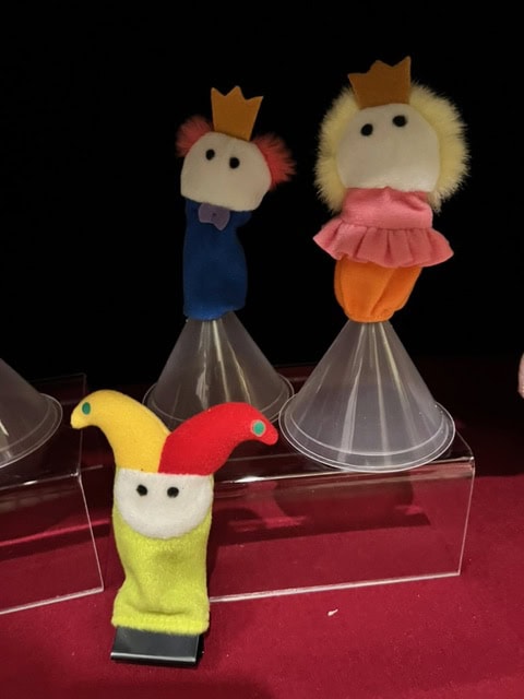 three finger puppets: two with crowns dressed as royalty, and one in a jester hat, are displayed on clear stands against a dark background.