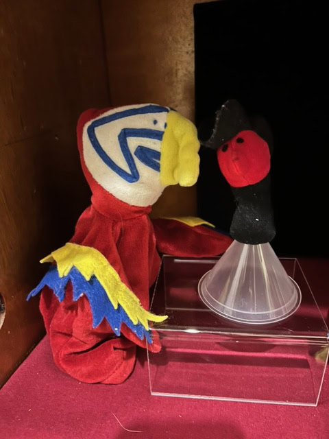 a red, yellow, and blue bird puppet faces a small, black and red figure with a cone shaped base on a clear display box against a wooden background.