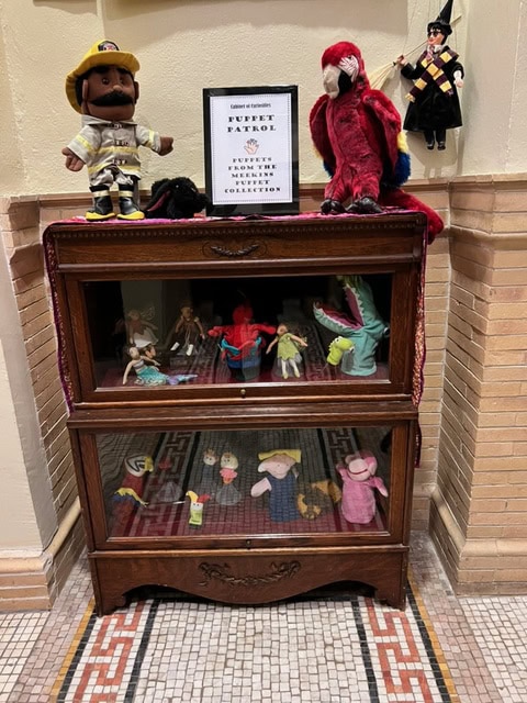 a wooden display case contains various puppets, with additional puppet figures and a sign reading 