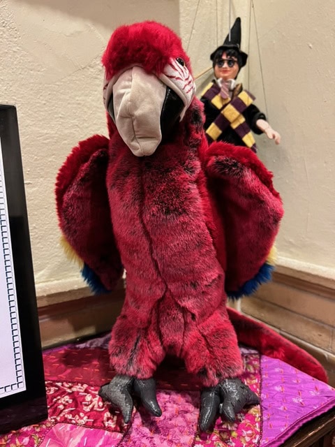a red plush parrot toy stands upright on a colorful fabric surface with a marionette doll hanging in the background.