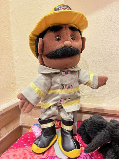 a plush firefighter doll with a yellow helmet, bushy mustache, tan uniform, and black boots stands upright on a colorful surface.