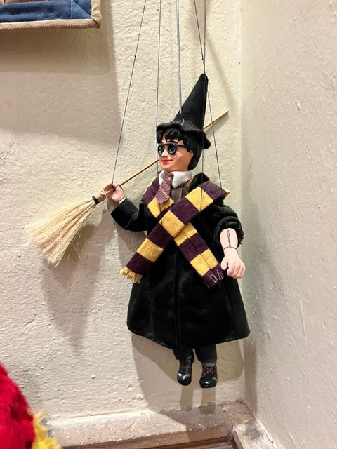 a marionette dressed as a wizard, wearing glasses, a black robe, a pointed hat, and a yellow and purple scarf, holding a broomstick, hangs against a corner wall.