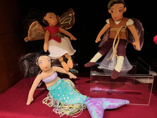 three fabric dolls: a mermaid with a blue green tail, a fairy with gold wings and a red dress, and a pirate with brown pants and a green headscarf, displayed on a red surface.