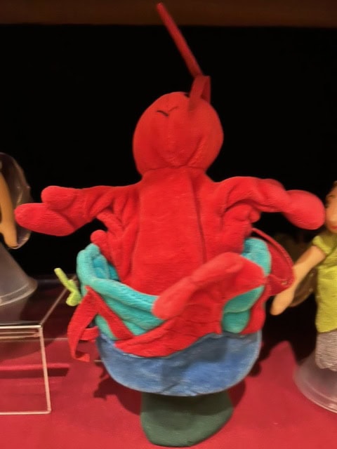 a red plush lobster toy sits upright in a blue and green cloth container, with its claws and antennae visible.
