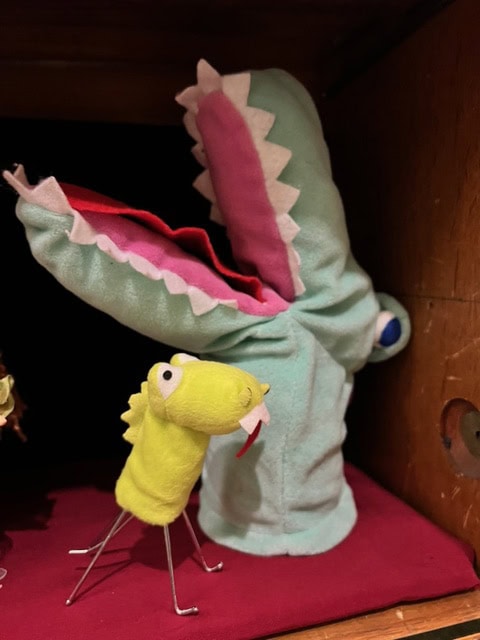 two handmade puppets: a large green crocodile with an open mouth and a smaller yellow creature with wire legs, displayed on a red cloth background.